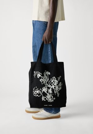 Black cotton tote bag with white floral graphic design, featuring sturdy handles and a rectangular shape. Worn by a person in blue jeans and shoes.