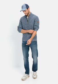 Fleece-Pullover in strukturiertem Blau mit einem Viertel-Reißverschluss und Logopatch am Ärmel. Kombiniert mit hellen Jeans und beigen Sneakers.