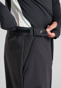 Svarta byxor med ett midjeband av kardborre och plastspänne. Materialet verkar slätt och flexibelt, med en strömlinjeformad design och snedställda sömmar.