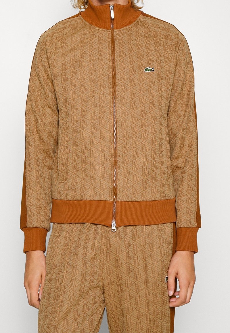 Braune Zip-Jacke mit geometrischem Muster, gerippten Bündchen und Saum sowie einem hohen Kragen. Verfügt über ein kleines grünes Logo auf der Brust.