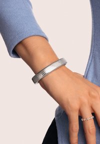 Bracciale rigido in argento con design scanalato, texture liscia e finitura lucida. Indossato su un polso insieme a un anello semplice.