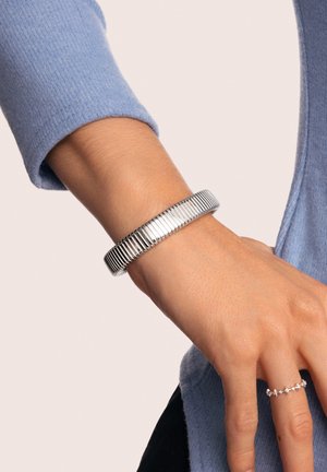 Bracciale rigido in argento con design scanalato, texture liscia e finitura lucida. Indossato su un polso insieme a un anello semplice.