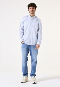 Lichtblauw overhemd met knopen en een subtiel patroon, gedragen met blauwe jeans en witte sneakers. Het overhemd heeft lange mouwen en een zachte textuur.