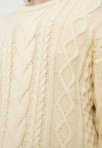 Πουλόβερ πλεκτό σε χρώμα κρέμα με υφασμένα μοτίβα καλωδίων, στρογγυλή λαιμόκοψη και ριμπ μανσέτες. Μαλακό, χοντρό ύφασμα.