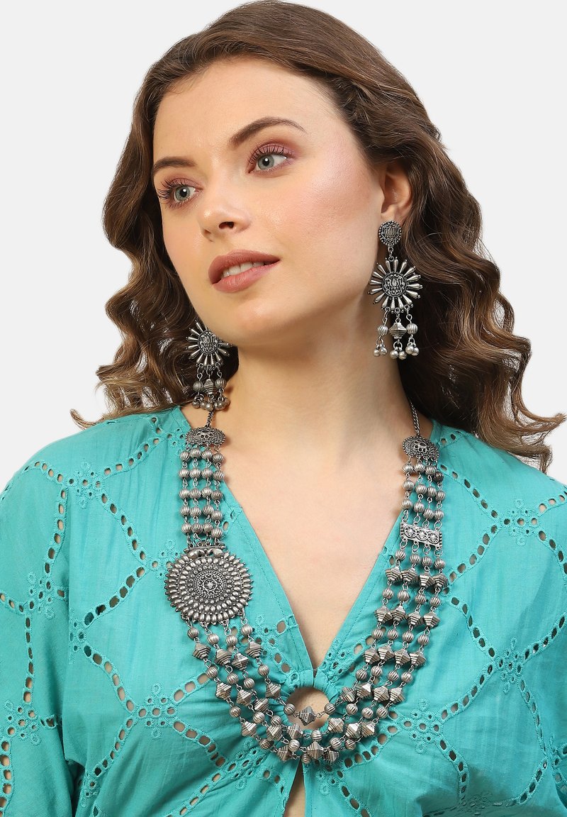 Collana multi-strato argento con medaglione circolare e dettagli intricati in perline. Il modello indossa una blusa turchese con motivi a taglio.