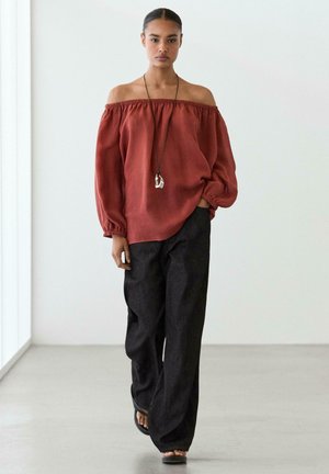 Woman wearing off-shoulder rust-colored blouse, loose black pants, pendant necklace, and black sandals standing in minimalist space.