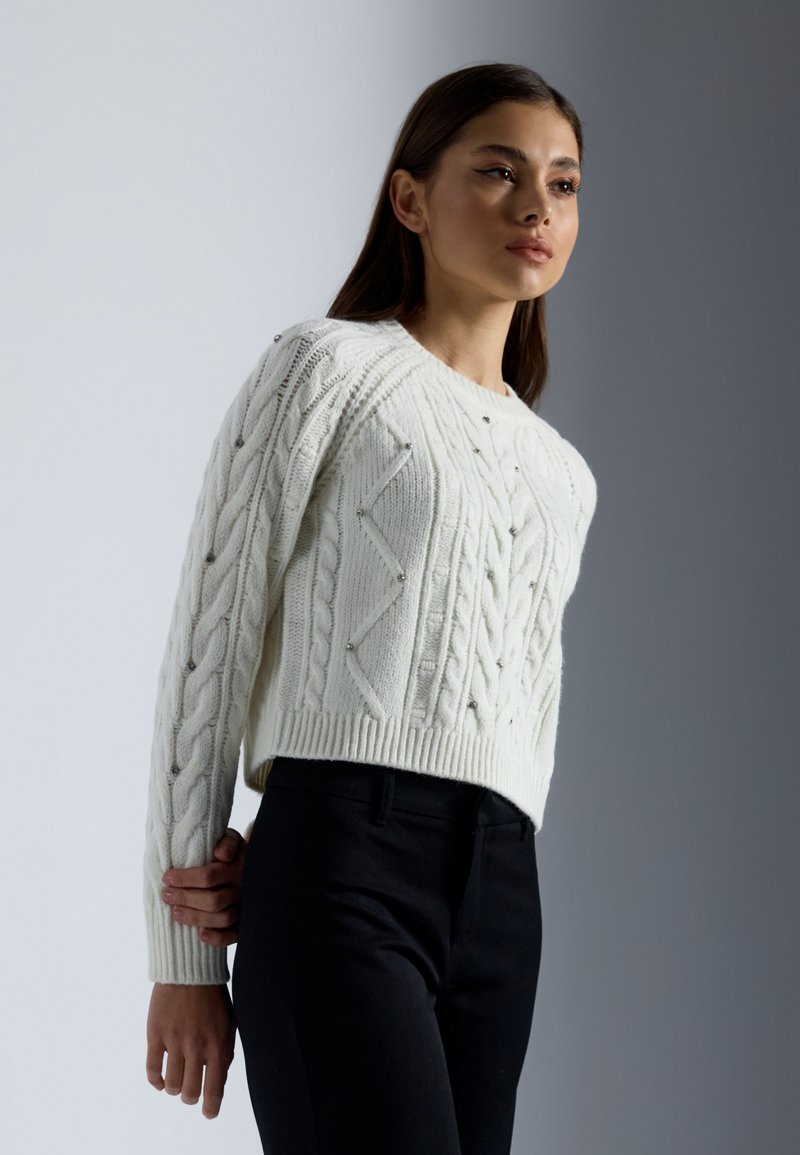 Maglione bianco lavorato a maglia con motivi a treccia testurizzati e piccole decorazioni, abbinato a pantaloni scuri, mostrato in profilo laterale.