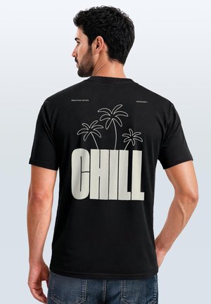 Mann mit dunklen Haaren, der ein schwarzes T-Shirt mit dem großen Wort "CHILL" und drei einfachen Palmenumrissen auf dem Rücken trägt, steht vor einem einfarbigen Hintergrund.