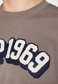 T-shirt en coton marron avec une grande application en velours bleu marine "1969" sur le devant, dotée d'un col rond côtelé. Texture de tissu lisse.