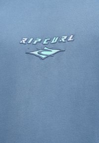 Maglietta a girocollo in cotone blu con logo "RIP CURL" in rilievo di colore azzurro e un elemento di design stilizzato al centro. Texture morbida, vestibilità casual.