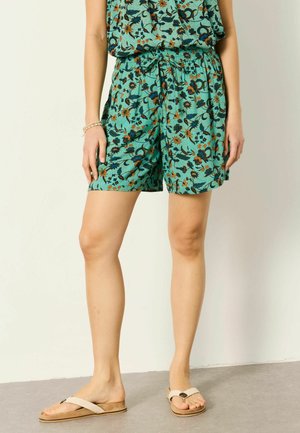 Femeie purtând șorturi cu imprimeu floral turcoaz, cu talie cu șnur, și sandale bej slip-on, stând în picioare lângă un perete simplu.