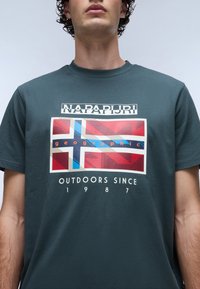 Tmavě zelené klasické bavlněné tričko s grafickým designem, který zobrazuje stylizovanou vlajku v červené, modré a krémové barvě, a nápis "NAFAPURI geographic OUTDOORS SINCE 1987."