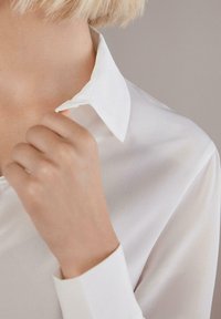Biała bluzka z długim rękawem, zaokrąglonym kołnierzykiem i gładką teksturą. Trzymana ręcznie w pobliżu kołnierzyka, ukazując miękki materiał i estetyczny design.
