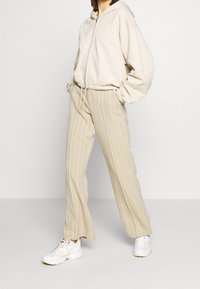 Personne portant un sweat à capuche beige zippé, un pantalon large beige à fines rayures et des baskets blanches, debout les mains dans les poches devant un fond uni.