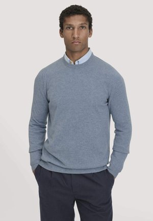 Pull bleu clair en maille douce, col rond et poignets côtelés. Porté par-dessus une chemise à col, avec une coupe décontractée.