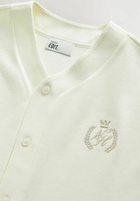 Lys gul bomulds cardigan med V-hals, med knapper og et broderet logo med en krone og laurbærkrans.