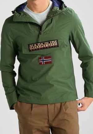 Mężczyzna w zielonej kurtce Napapijri z przednią kieszenią na zamek i naszywką z norweską flagą, zestawionej z brązowymi spodniami.