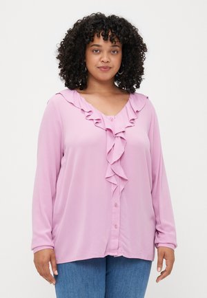 Blouse rose clair avec un col volanté et des manches longues. Présente une fermeture à boutons sur le devant et une texture douce.