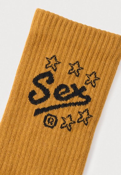 Κίτρινη ριγωτή κάλτσα με μαύρα γράμματα που γράφουν «Sex» και πέντε μαύρα αστέρια. Η υφή είναι μαλακή και πλεκτή με ελαστική εφαρμογή.
