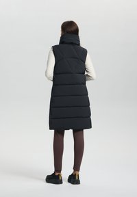 Veste sans manches noire avec col haut et coutures verticales, design superposé, portée sur un haut clair, associée à des leggings marron et des chaussures noires.