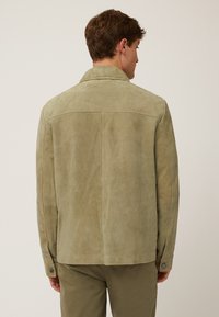Veste en daim de couleur olive clair avec une coupe droite, un col pointu, des poignets boutonnés et des coutures minimales visibles. Texture lisse.
