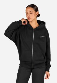 Fekete cipzáras kapucnis pulóver, puha anyagból készült, nagy kapucnival, elülső zsebekkel és bal mellen található írásos logóval. Laza fazon.