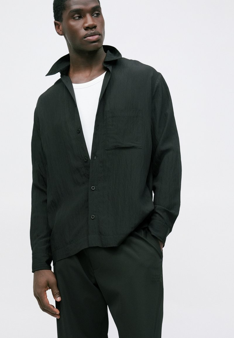 Ung mand med mørk hud iført en sort skjorte med knapper over en hvid t-shirt og sorte bukser foran en ensfarvet lys baggrund.