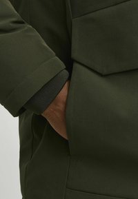 Tumedat rohelised jakikangas, millel on sile tekstuur, ribitud mansett ja küljetasku. Lihtne disain, minimaalse aktsentiidiga.