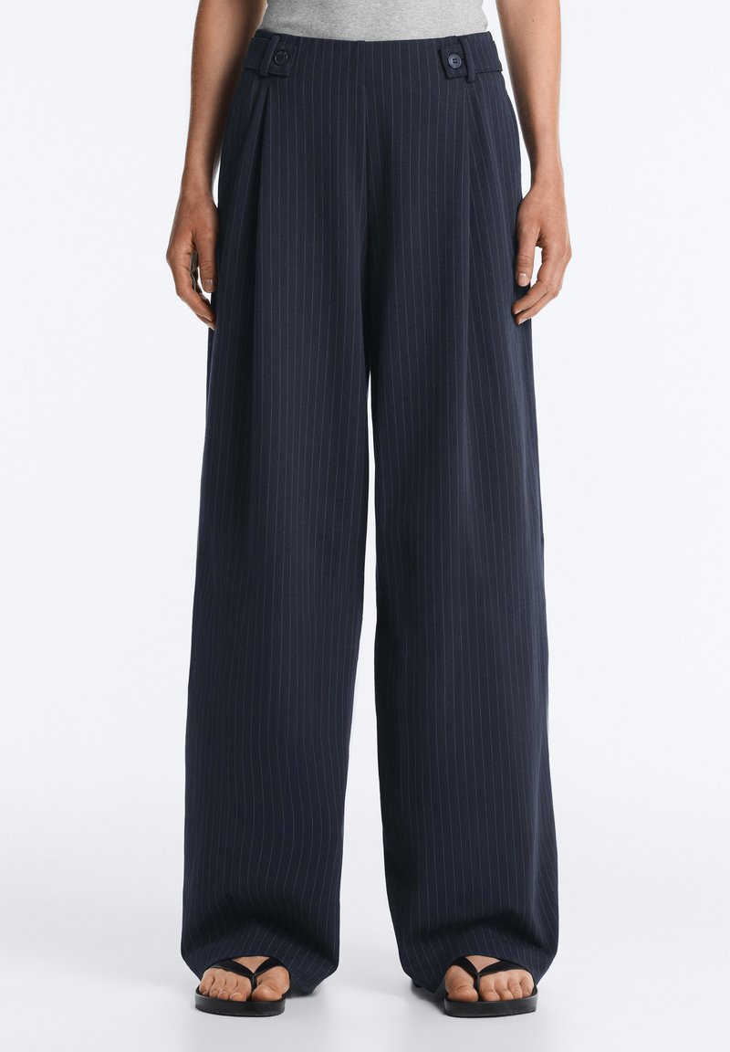 Pantalon large à rayures fines bleu marine avec des détails de boutons à la taille, porté avec un haut gris et des sandales noires à bout ouvert.