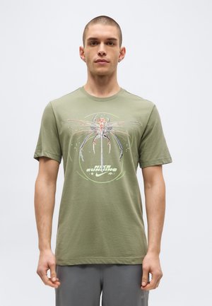 Vyras dėvi alyvuogių žalsvos spalvos „Nike Running“ marškinėlį su abstrakčiu vabzdžio piešiniu, stovi prieš paprastą šviesų foną.