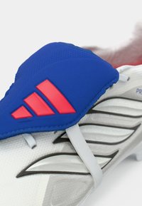 Λευκό ποδοσφαιρικό παπούτσι με μπλε επάνω μέρος, που διαθέτει κόκκινες λεπτομέρειες και υφή. Περιλαμβάνει γκρίζα διακοσμητικά και ελαστικές ιμάντες για καλύτερη εφαρμογή.