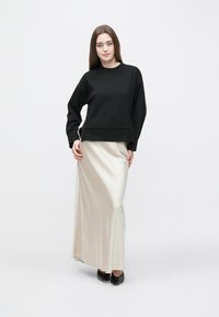 Μαύρο φαρδύ φούτερ από απαλό ύφασμα, σε συνδυασμό με μακριά, κρεμ σατέν φούστα που ρέει μέχρι το πάτωμα, φορεμένο με μαύρα παπούτσια.