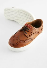 Chaussure en cuir marron avec des perforations et une finition lisse. Semelle en caoutchouc blanc avec grip texturé. Lacets noirs et rouges.