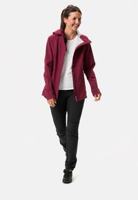 Giacca impermeabile bordeaux con cappuccio, chiusura a zip e maniche regolabili, abbinata a pantaloni neri e scarpe nere, indossata da un modello.