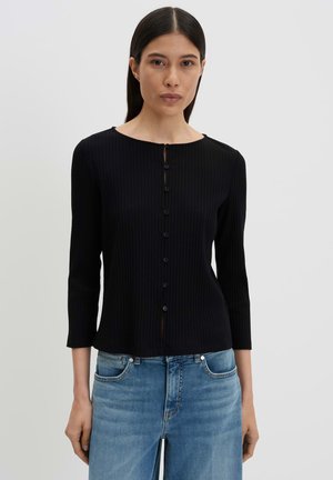 Vrouw met steil donker haar draagt een aansluitende zwarte geribbelde cardigan met knopen en lichtblauwe spijkerbroek tegen een effen grijze achtergrond.