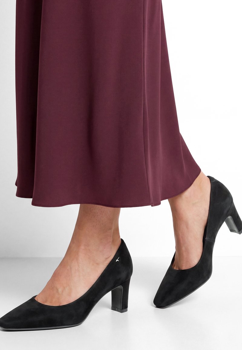 Femme portant des chaussures en daim noir à talons moyens et une longue jupe bordeaux, debout sur un sol blanc.
