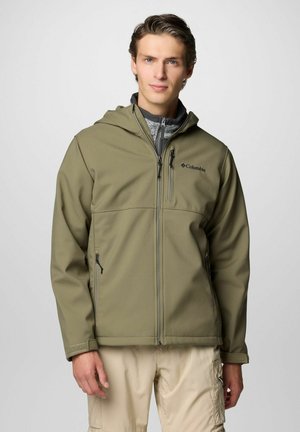 Giacca softshell verde oliva con cappuccio, zip frontale e due tasche laterali. Presenta una leggera fodera grigia e un logo discreto sul petto.