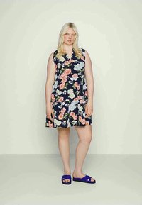 Jovem mulher com cabelo loiro longo, vestindo um vestido sem mangas azul-marinho com estampado floral em rosa, branco e azul, e sandálias azuis, de pé contra uma parede clara.