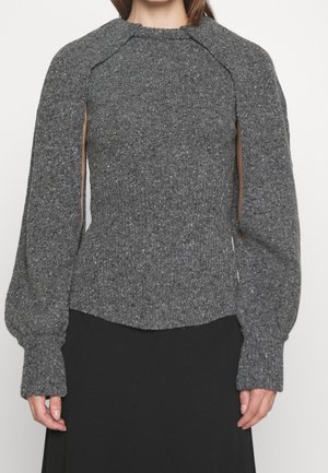 Frau trägt einen grauen, strukturierten Strickpullover mit offenen Ärmelschlitzen von der Schulter bis zum Ärmelbündchen, kombiniert mit einem schwarzen Rock.