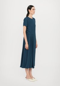 Kleid knielang in dunklem Teal mit kurzen Ärmeln, das einen taillierten Oberteil, einen gefältelten Rock und eine subtile Textur aufweist. Trägt weiße Mules.