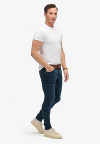 Camiseta blanca de cuello redondo combinada con jeans ajustados azul oscuro y zapatillas beige claras. El cinturón es marrón con una hebilla clásica.