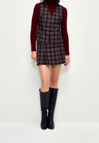 Rutigt väst med knappar, parat med en matchande kjol. Turtleneck i vinröd och knähöga svarta läderstövlar. Texturerade tyger visas.
