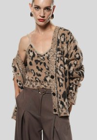 Cardigan beige con stampa leopardata e dettagli neri, abbinato a una canotta coordinata. Pantaloni marroni a vita alta con una cintura texturizzata.