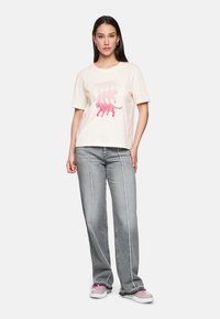 Eine Frau steht und trägt ein cremefarbenes T-Shirt mit drei rosa Leopardenmustern, graue weit geschnittene Jeans mit vertikalen Nähten und pinke Turnschuhe.