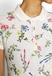 Poloshirt in hellem Creme mit einem floralen Muster, das bestickte Blumen in Pink, Blau, Gelb und Grün zeigt; kurze Ärmel und Knopfleiste.
