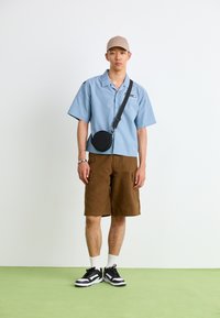 Lyseblå kortermet skjorte, brune cargo shorts, svart rund krysseveske, svart og hvit sneakers, og en beige caps.