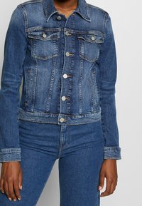 Blå denimjacka med en kortad design, klassisk krage, bröstfickor och silverfärgade metallknappar. Har en urtvättad textur och kontrasterande sömmar.