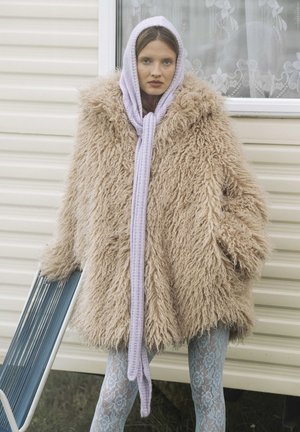 Zachte beige bontjas met grote capuchon, gecombineerd met een lila gebreide sjaal. Het model draagt lichtblauwe legging met kantmotief.