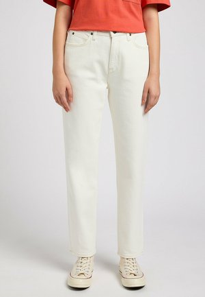 Osoba ubrana w kremowe, proste dżinsy, białe sznurowane trampki i pomarańczowy krótki top na gładkim tle.