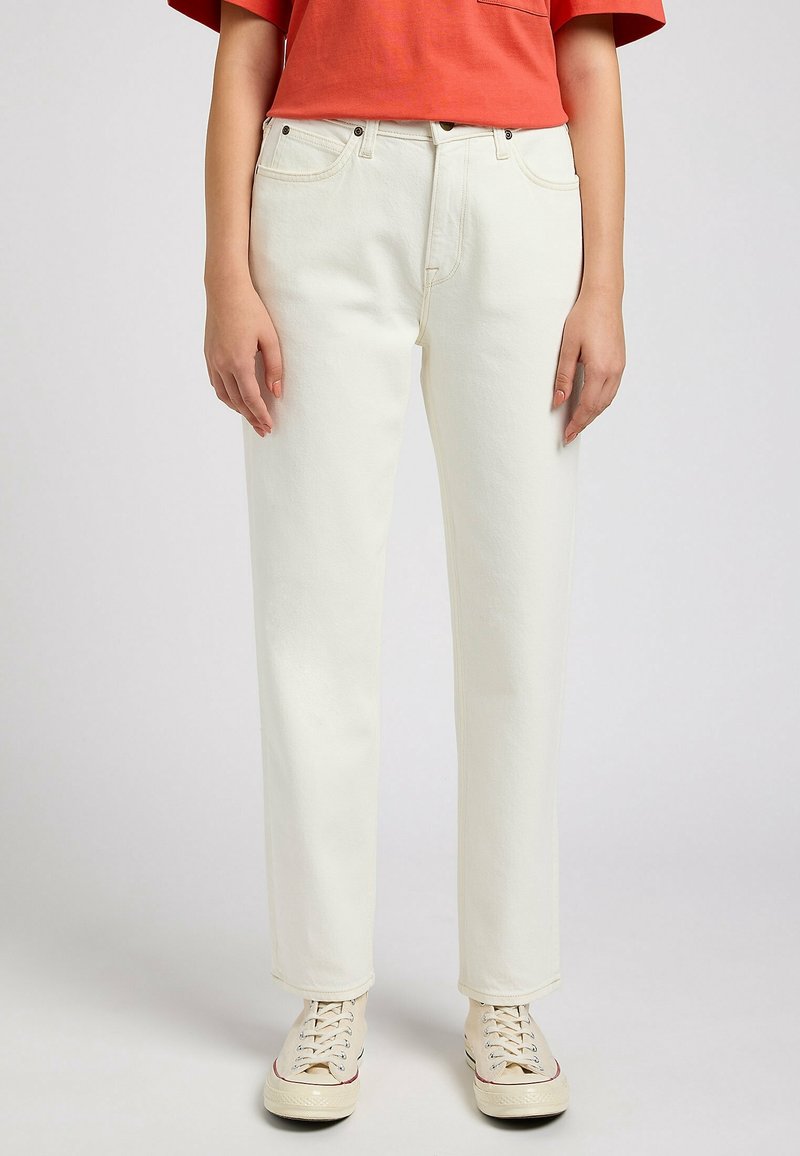 Personne portant un jean droit blanc cassé, des baskets blanches à lacets et un haut orange à manches courtes sur un fond uni.